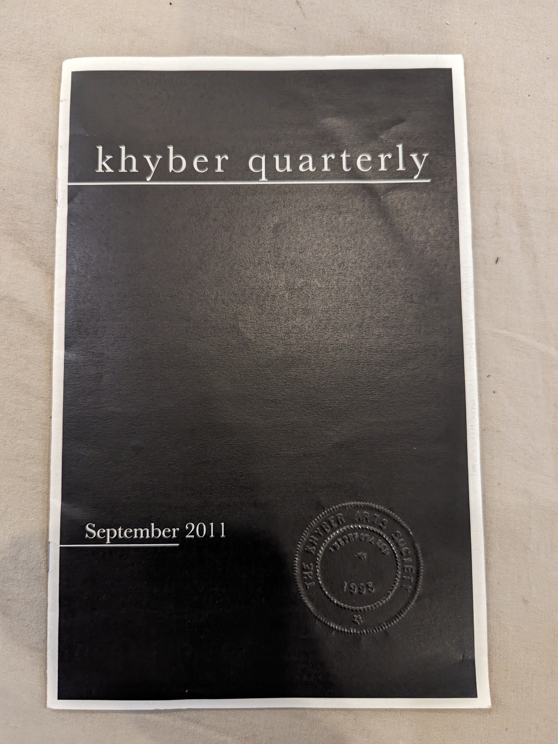 Black half page zine with a white border. A Khyber seal is on the bottom right corner of the zine